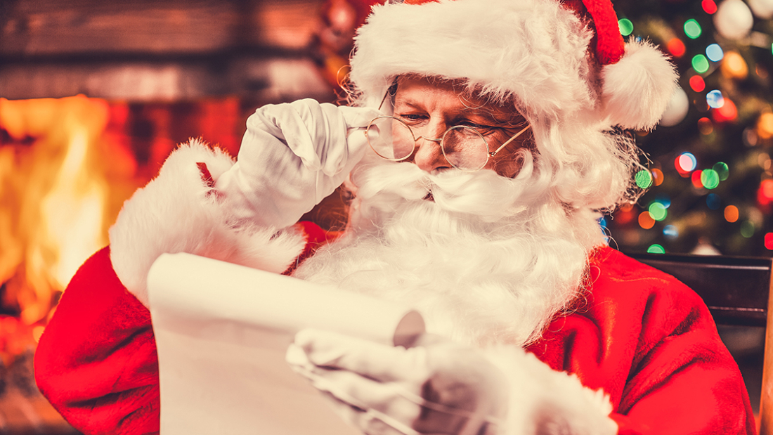 Traditional Santa Claus sitting at his chair and reading a letter with fireplace and Christmas Tree in the background